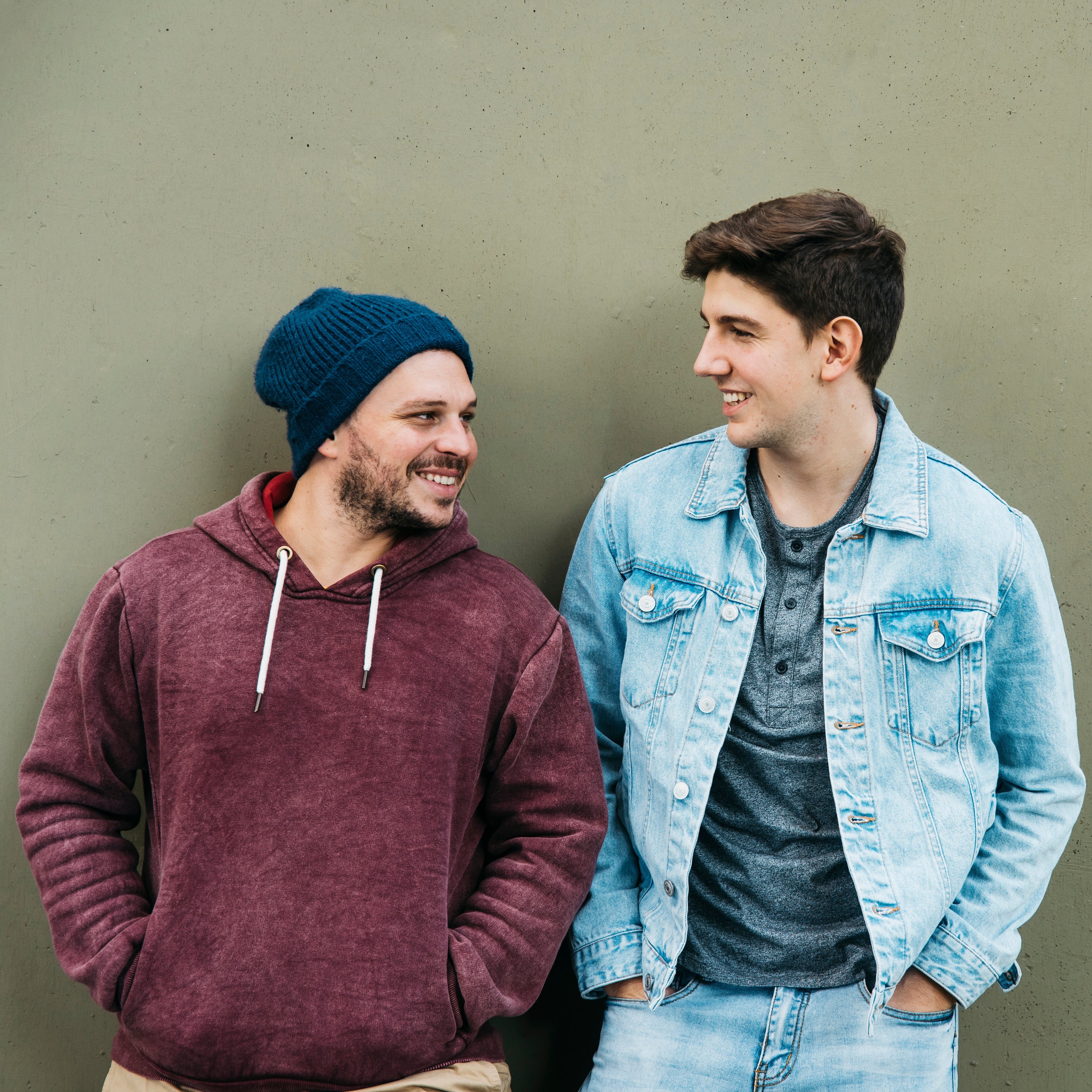 Two men standing against a plain wall, one wearing a maroon hoodie and blue beanie, the other in a denim jacket and gray shirt.